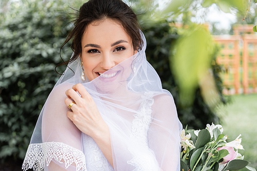 Cheerful bride holding white veil near bouquet outdoors