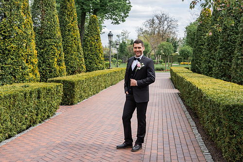 Cheerful groom holding hand in pocket of pants in park