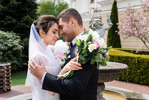 Side view of smiling groom hugging bride with bouquet near blurred fountain