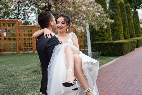 Groom holding on hands smiling bride in white wedding dress in park