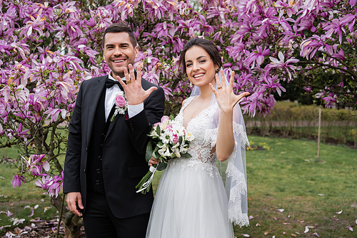 Smiling bride and groom showing rings near magnolia trees