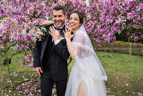 Cheerful bride sticking out tongue near groom showing ring in park