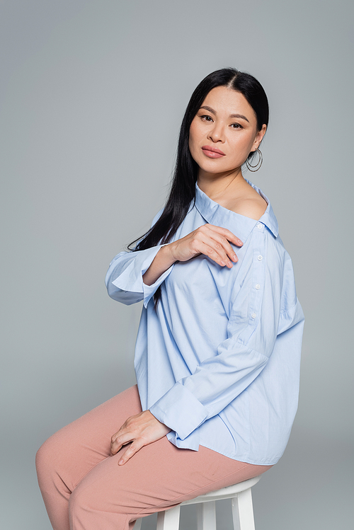 Asian woman in blouse with naked shoulder sitting on chair isolated on grey