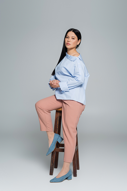 Full length of stylish asian woman posing near chair on grey background
