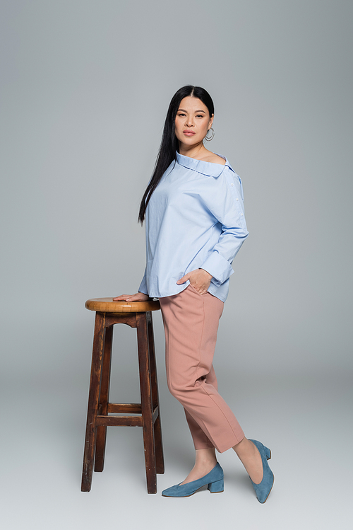 Full length of stylish asian woman holding hand in pocket of pants near chair on grey background