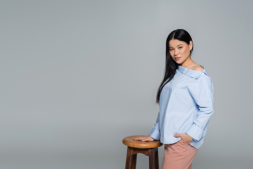 Asian model in blue blouse posing near chair isolated on grey