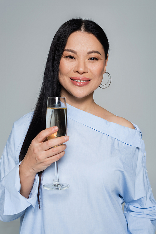 Smiling asian woman in blouse holding glass of champagne isolated on grey