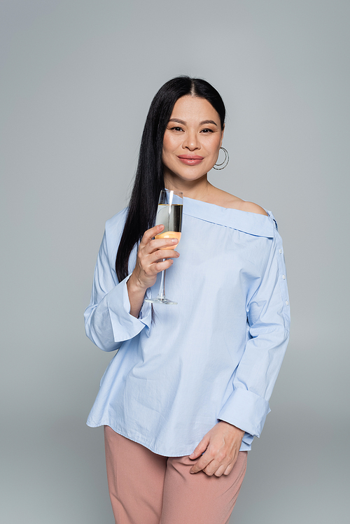 Stylish asian woman holding glass of champagne and smiling isolated on grey