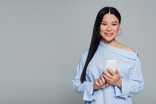 Positive asian woman in blouse using mobile phone isolated on grey