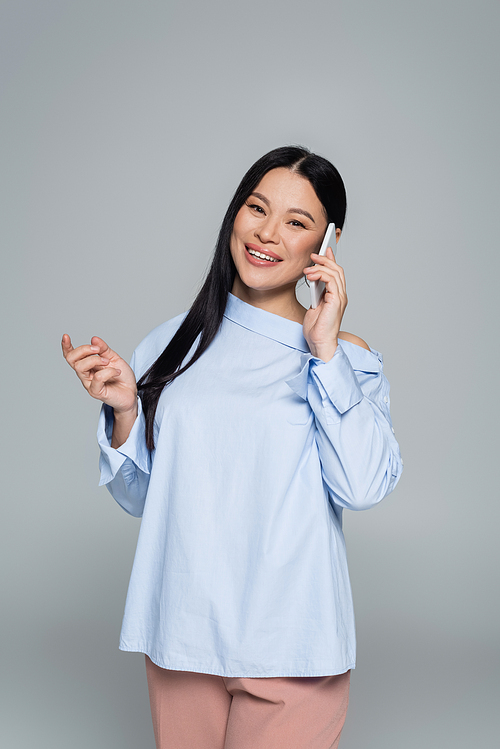 Smiling asian model in blouse talking on smartphone isolated on grey