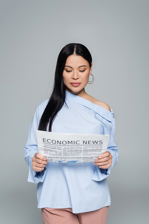 Stylish brunette asian woman reading economic news isolated on grey