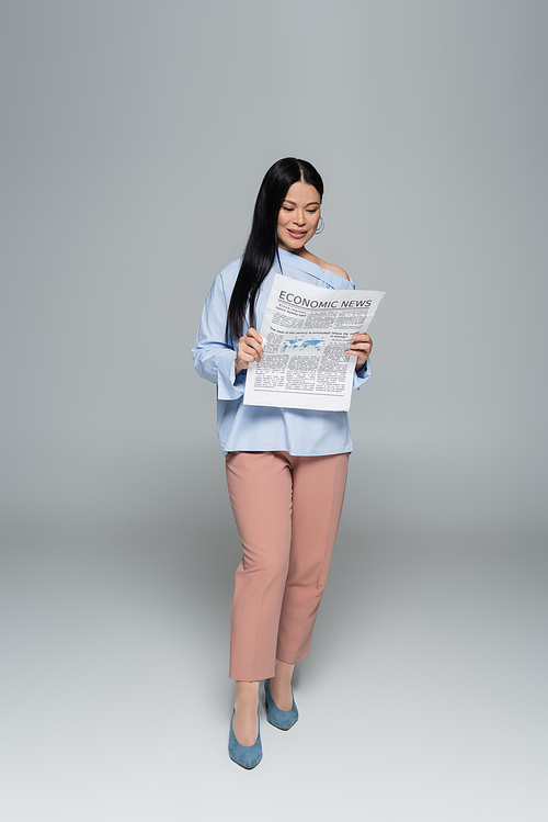 Full length of smiling asian woman reading economic newspaper on grey background