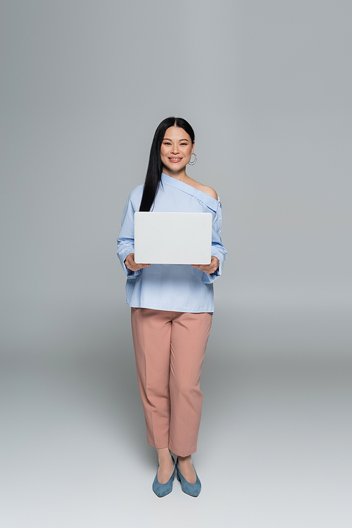 Positive and stylish asian woman holding laptop on grey background