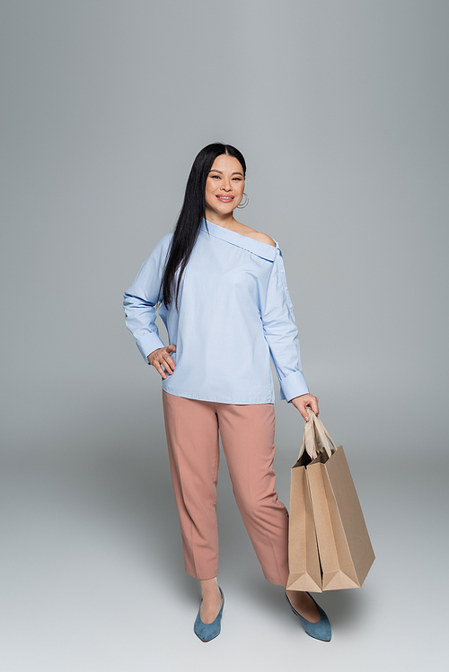 Full length of smiling asian woman holding shopping bags on grey background