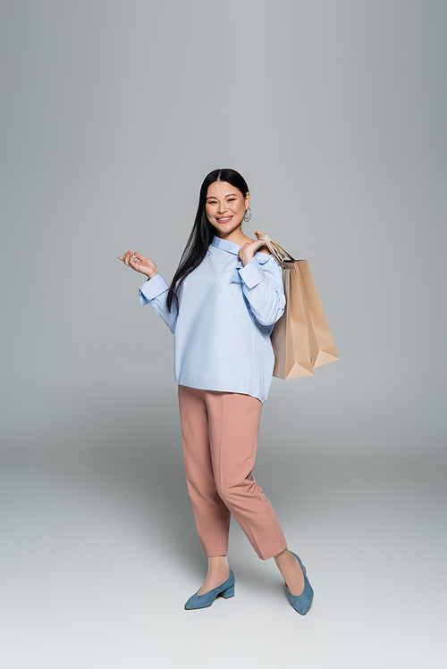 Positive asian model holding shopping bags and pointing with finger on grey background