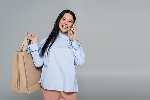 Smiling asian woman holding shopping bags and talking on smartphone isolated on grey