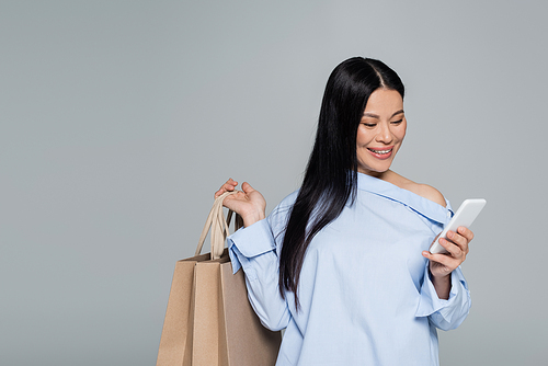 Positive asian woman using smartphone and holding shopping bags isolated on grey