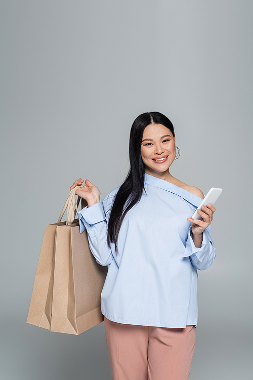 Smiling asian woman holding smartphone and shopping bags isolated on grey