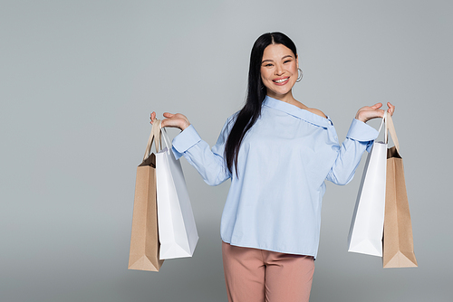 Cheerful asian woman in blouse holding purchases isolated on grey