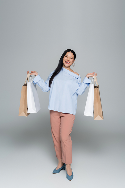 Full length of smiling positive asian woman holding shopping bags on grey background