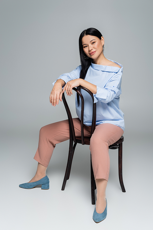 Smiling asian woman  while sitting on chair on grey background