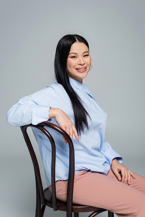 Positive asian woman in blouse sitting on chair and  isolated on grey