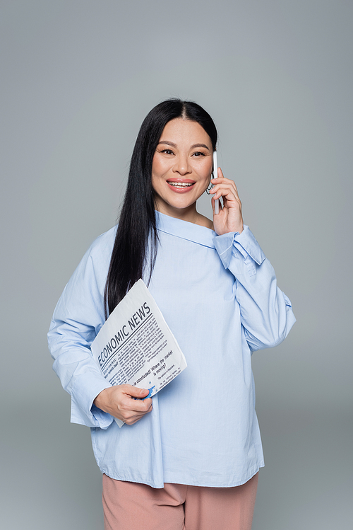 Smiling asian woman holding economic newspaper and talking on smartphone isolated on grey