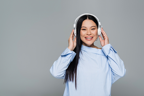 Cheerful asian woman in blouse listening music in headphones isolated on grey