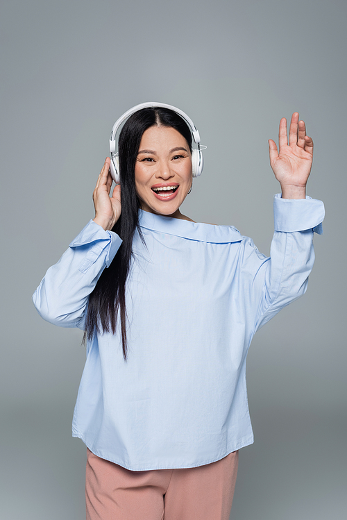 Positive asian woman in headphones waving hand isolated on grey