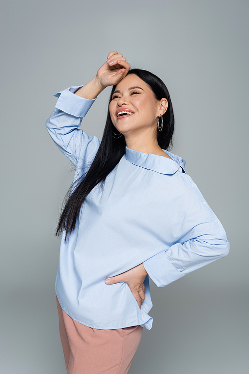 Positive asian woman in blue blouse holding hand on hip isolated on grey