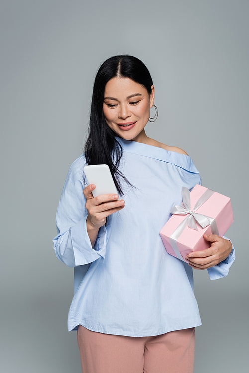 Positive asian woman using smartphone and holding gift isolated on grey