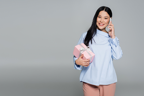 Cheerful asian woman holding present and talking on smartphone isolated on grey