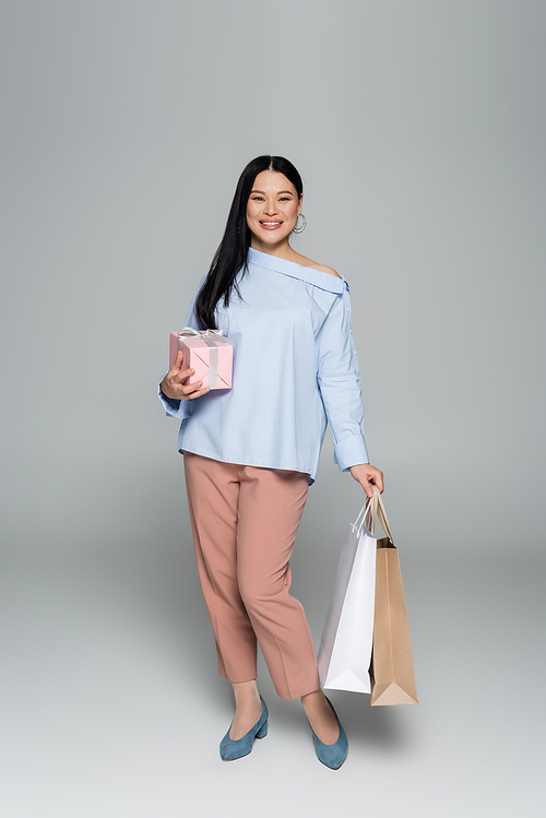 Cheerful asian woman holding gift and shopping bags on grey background