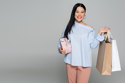 Smiling asian woman holding shopping bags and present isolated on grey