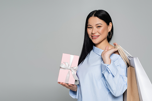Positive asian model holding purchases and gift isolated on grey