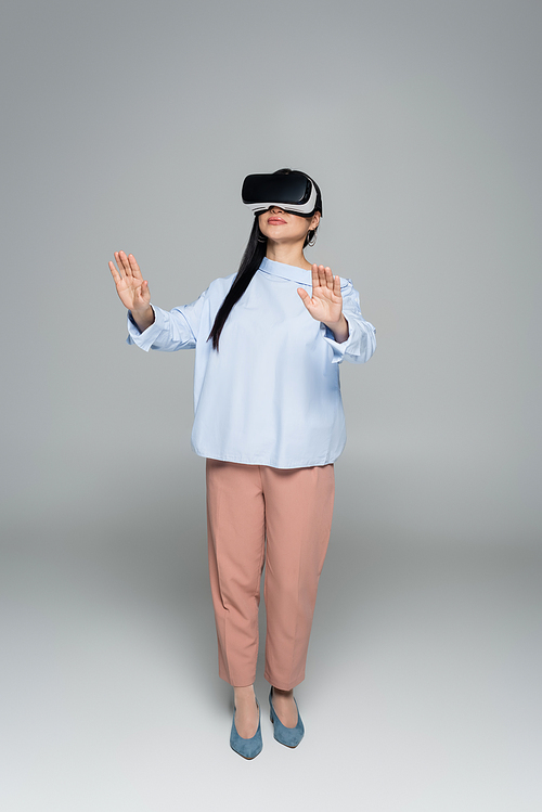 Full length of stylish woman in virtual reality headset on grey background
