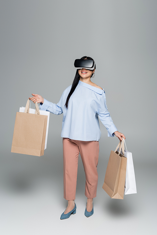 Smiling woman in vr headset holding shopping bags on grey background