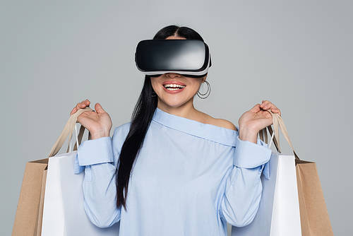 Cheerful woman in vr headset holding shopping bags isolated on grey