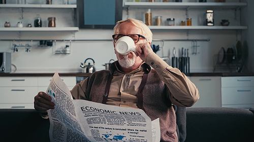 Senior man in eyeglasses holding newspaper while drinking tea at home