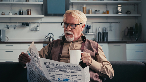 Senior man in eyeglasses reading newspaper and holding cup of tea at home