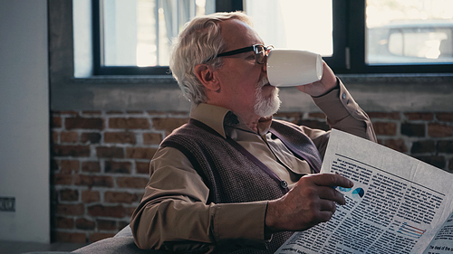 bearded senior man in eyeglasses holding newspaper while drinking tea