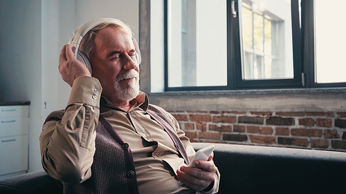 pleased and senior man in wireless headphones listening music and holding smartphone at home