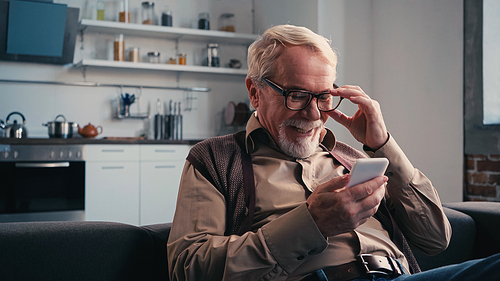 happy senior man adjusting eyeglasses and using cellphone at home
