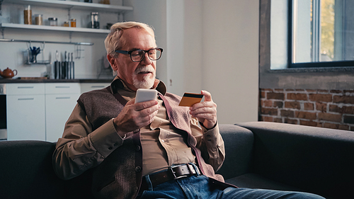 pensioner in eyeglasses with credit card holding mobile phone while shopping online at home