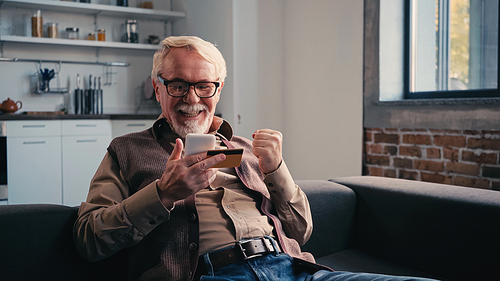 happy pensioner with credit card holding mobile phone while shopping online at home
