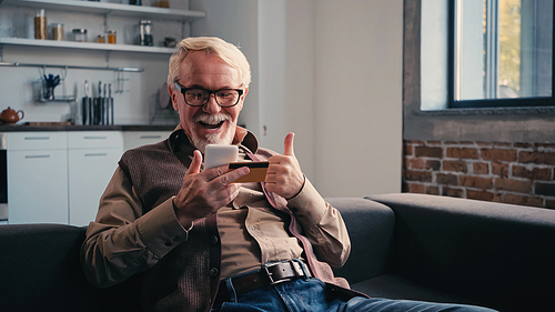 happy pensioner with credit card holding mobile phone while showing thumb up at home