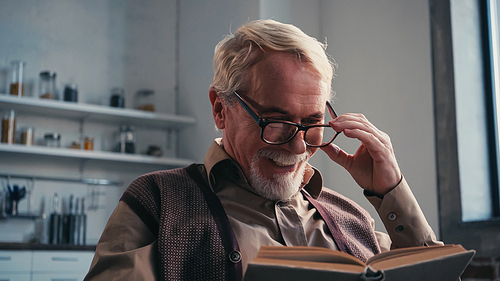 positive senior man adjusting eyeglasses and reading book at home