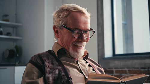 positive senior man in eyeglasses reading book at home