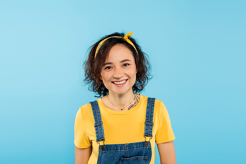pretty woman in headband and denim overalls smiling at camera isolated on blue
