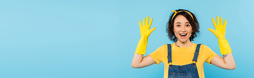 amazed housewife showing hands in yellow rubber gloves isolated on blue, banner